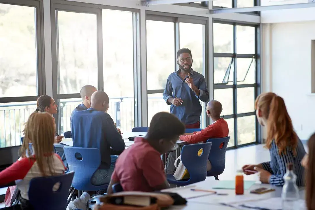 A group of people sitting around tables in a classroom listen to a presenter