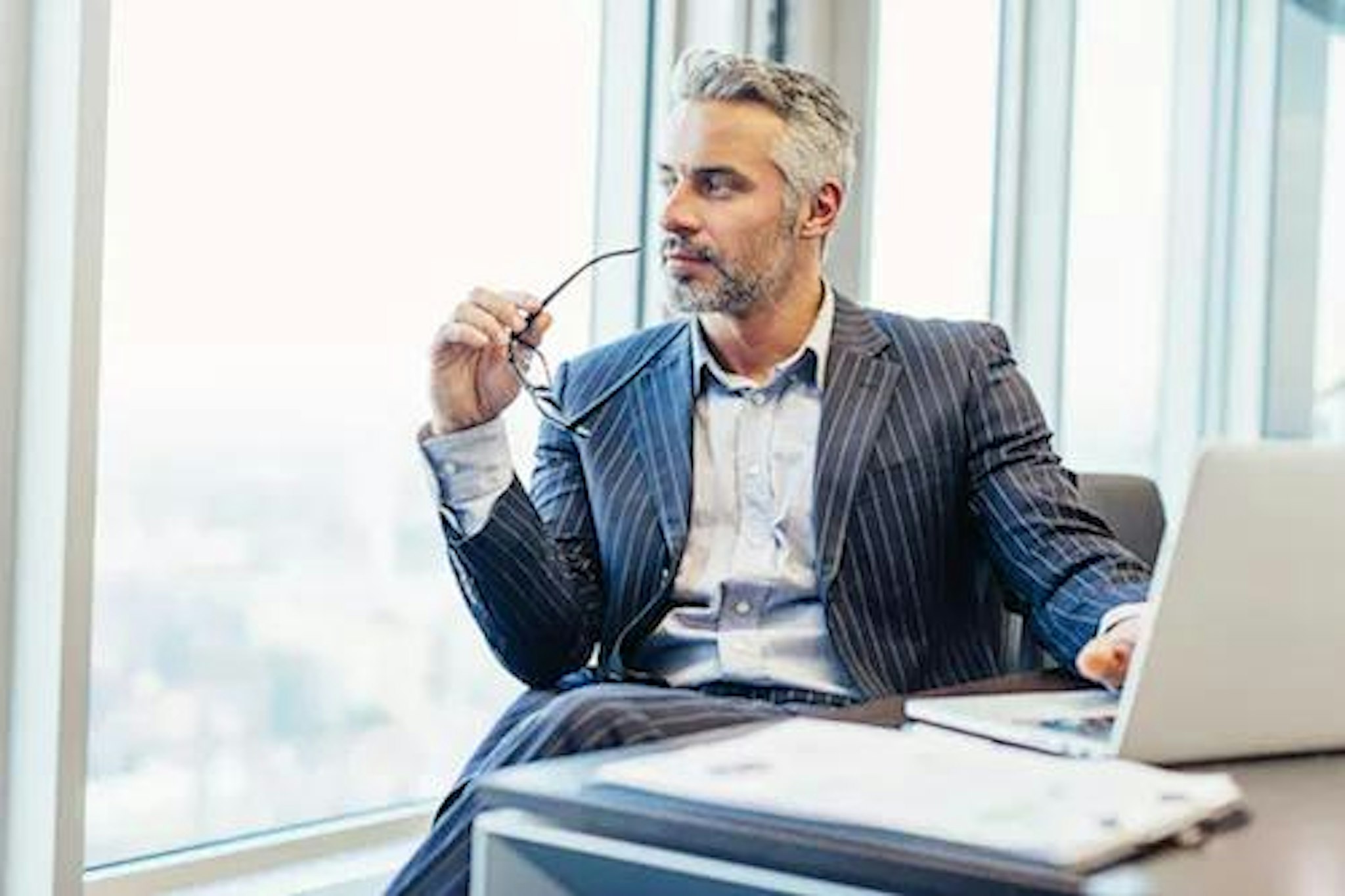 Person sits in chair holding glasses and looking out his office window while considering why organizations split the CEO and chairman of the board role