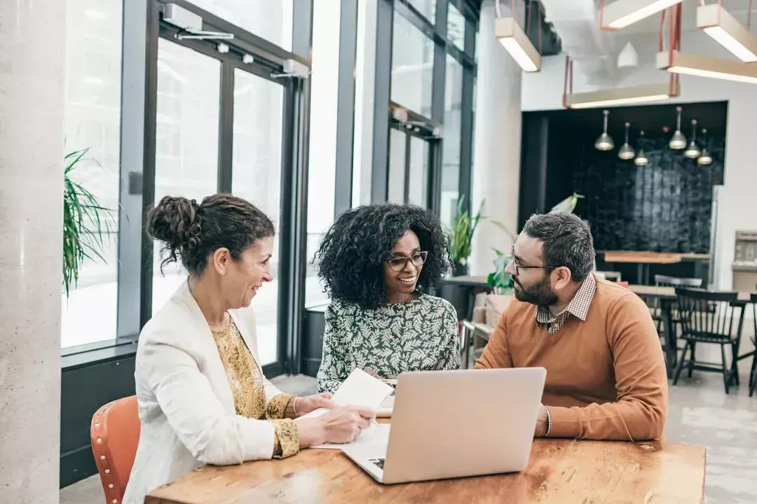 Three people sit at a table in an office around a laptop, having a conversation